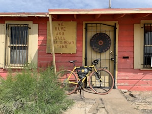 A rustic building with weathered red paint and yellow trim features a sign advertising railroad ties for sale. In front of the building is a yellow bicycle with gear attached, leaning against the doorway. A patch of green shrubbery grows in the foreground.