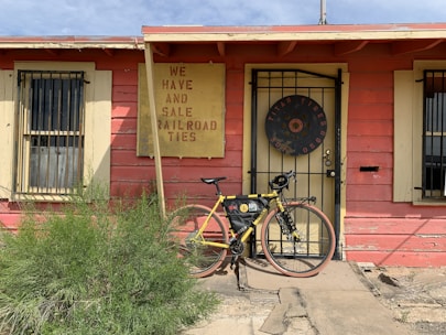 A rustic building with weathered red paint and yellow trim features a sign advertising railroad ties for sale. In front of the building is a yellow bicycle with gear attached, leaning against the doorway. A patch of green shrubbery grows in the foreground.