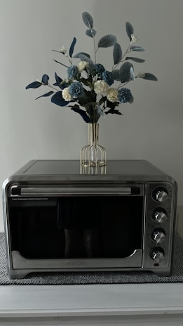 A countertop oven with a sleek metallic finish is placed on a textured mat. Above the oven, there is a decorative arrangement consisting of blue and white artificial flowers with green leaves in a transparent vase.