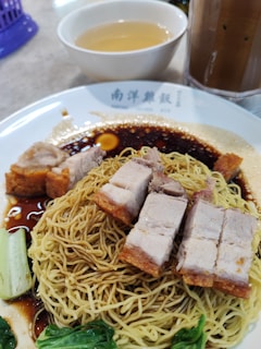 A plate of crispy roast pork with yellow noodles served on a white plate, accompanied by soy-based sauce and green vegetables. In the background, there is a bowl of clear soup and a tall glass of iced drink.