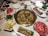 A table set for a hot pot meal. The center of the table features a divided pot with boiling broth and various vegetables. Surrounding the pot are plates of raw beef slices, meatballs, leafy greens, and condiments. There are also cups filled with drinks, bowls, chopsticks, and different plates and utensils.