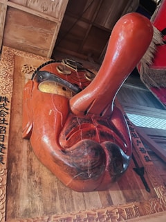 A large wooden mask featuring exaggerated facial features, prominently a long nose and thick mustache. It is painted in vibrant orange and black with gold accents around the eyes. The mask is mounted on a wooden surface adorned with intricate carvings and Japanese characters.
