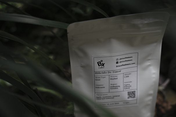A white coffee bag labeled 'Bix Coffee' sits partially obscured by green foliage. The label provides information about the coffee, including its origin from Indonesia, roast type as Omni, and process as washed. A QR code and contact information are also visible on the package.
