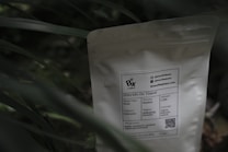 A white coffee bag labeled 'Bix Coffee' sits partially obscured by green foliage. The label provides information about the coffee, including its origin from Indonesia, roast type as Omni, and process as washed. A QR code and contact information are also visible on the package.