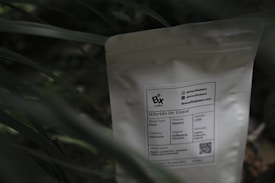 A white coffee bag labeled 'Bix Coffee' sits partially obscured by green foliage. The label provides information about the coffee, including its origin from Indonesia, roast type as Omni, and process as washed. A QR code and contact information are also visible on the package.