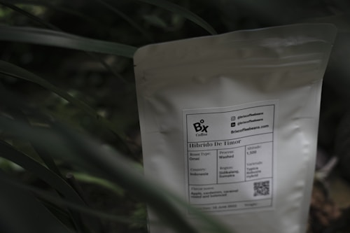 A white coffee bag labeled 'Bix Coffee' sits partially obscured by green foliage. The label provides information about the coffee, including its origin from Indonesia, roast type as Omni, and process as washed. A QR code and contact information are also visible on the package.