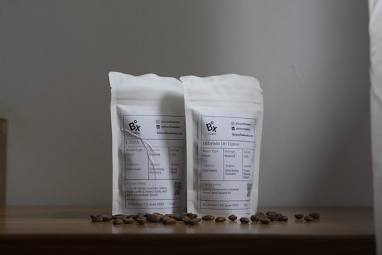 A simple wooden surface holds two white, sealed pouches labeled with information related to coffee. Scattered coffee beans complement the minimalist style of the composition. The background is plain, enhancing the focus on the coffee packages.
