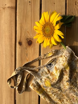 A bright yellow sunflower lies on a wooden surface beside a rustic, patterned tote bag. The wooden planks have a light, natural finish, and there is a label sewn into the bag.