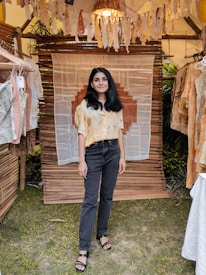 A person stands in a rustic setting with hanging textile decor and various shirts on display. The backdrop includes bamboo structures and greenery, creating a warm, handcrafted ambiance.