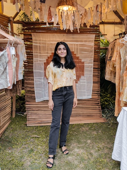 A person stands in a rustic setting with hanging textile decor and various shirts on display. The backdrop includes bamboo structures and greenery, creating a warm, handcrafted ambiance.