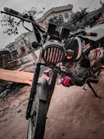 A custom motorcycle with a rugged appearance is parked on a dirt path. The bike is designed with a matte black paint job, featuring a branded logo of 'Cafe Racer'. It is equipped with dual front suspensions, a circular headlamp with grill protection, and handlebars extending outward. The background includes residential buildings and some trees.