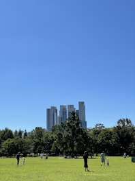 a group of people walking in a park with trees and tall buildings in the background