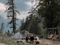 A camping scene with a tent set up among tall pine trees, with two people seated at a wooden table overlooking a mountainous and forested landscape. There are chairs and a swing nearby, and the sky is overcast, suggesting a calm, serene environment.