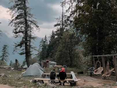 A cozy camping setup with a tent, campfire, and comfortable chairs under tall pine trees.