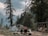 A camping scene with a tent set up among tall pine trees, with two people seated at a wooden table overlooking a mountainous and forested landscape. There are chairs and a swing nearby, and the sky is overcast, suggesting a calm, serene environment.