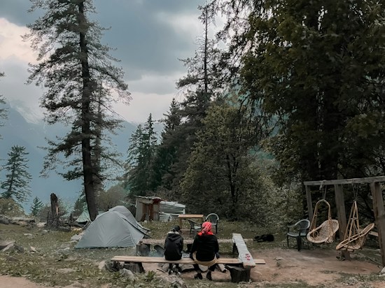 A camping scene with a tent set up among tall pine trees, with two people seated at a wooden table overlooking a mountainous and forested landscape. There are chairs and a swing nearby, and the sky is overcast, suggesting a calm, serene environment.