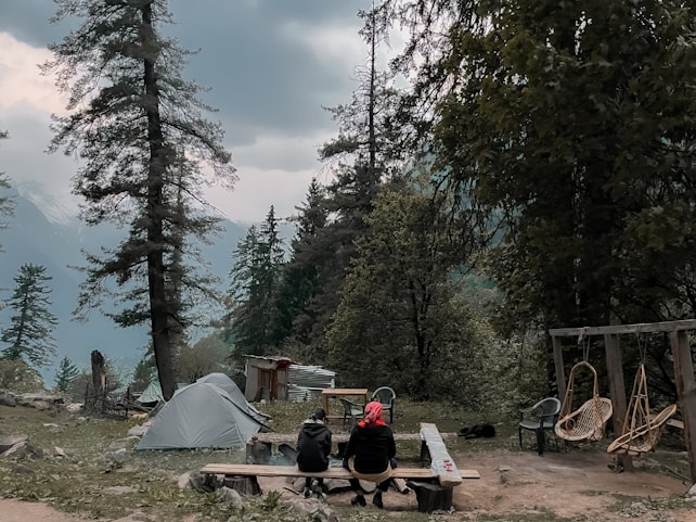 A camping scene with a tent set up among tall pine trees, with two people seated at a wooden table overlooking a mountainous and forested landscape. There are chairs and a swing nearby, and the sky is overcast, suggesting a calm, serene environment.