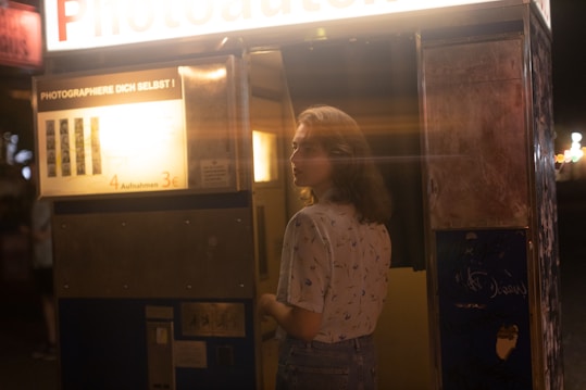 A woman stands in front of an old-fashioned photo booth at night. The booth is illuminated, casting a warm glow against the dark surroundings. The light reveals a partially obscured price list and photo instructions in a foreign language on the outside of the booth. The woman's expression is neutral, and she is dressed in a light blouse with subtle patterns.
