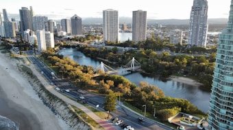 A vibrant cityscape with tall skyscrapers lining a scenic coastal area. A river winds through the city, crossed by an elegant suspension bridge. The shoreline features a sandy beach, while a road runs parallel to it, bordered by green trees and parks.