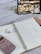 A detailed view of a study setup on a bed, including a tablet displaying study-related images, a spiral notebook open to pages filled with mathematical calculations, a pink calculator, and a set of wireless earbuds in their case. The notebook is placed on a clipboard, and a pen is seen nearby.