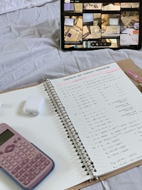 A detailed view of a study setup on a bed, including a tablet displaying study-related images, a spiral notebook open to pages filled with mathematical calculations, a pink calculator, and a set of wireless earbuds in their case. The notebook is placed on a clipboard, and a pen is seen nearby.