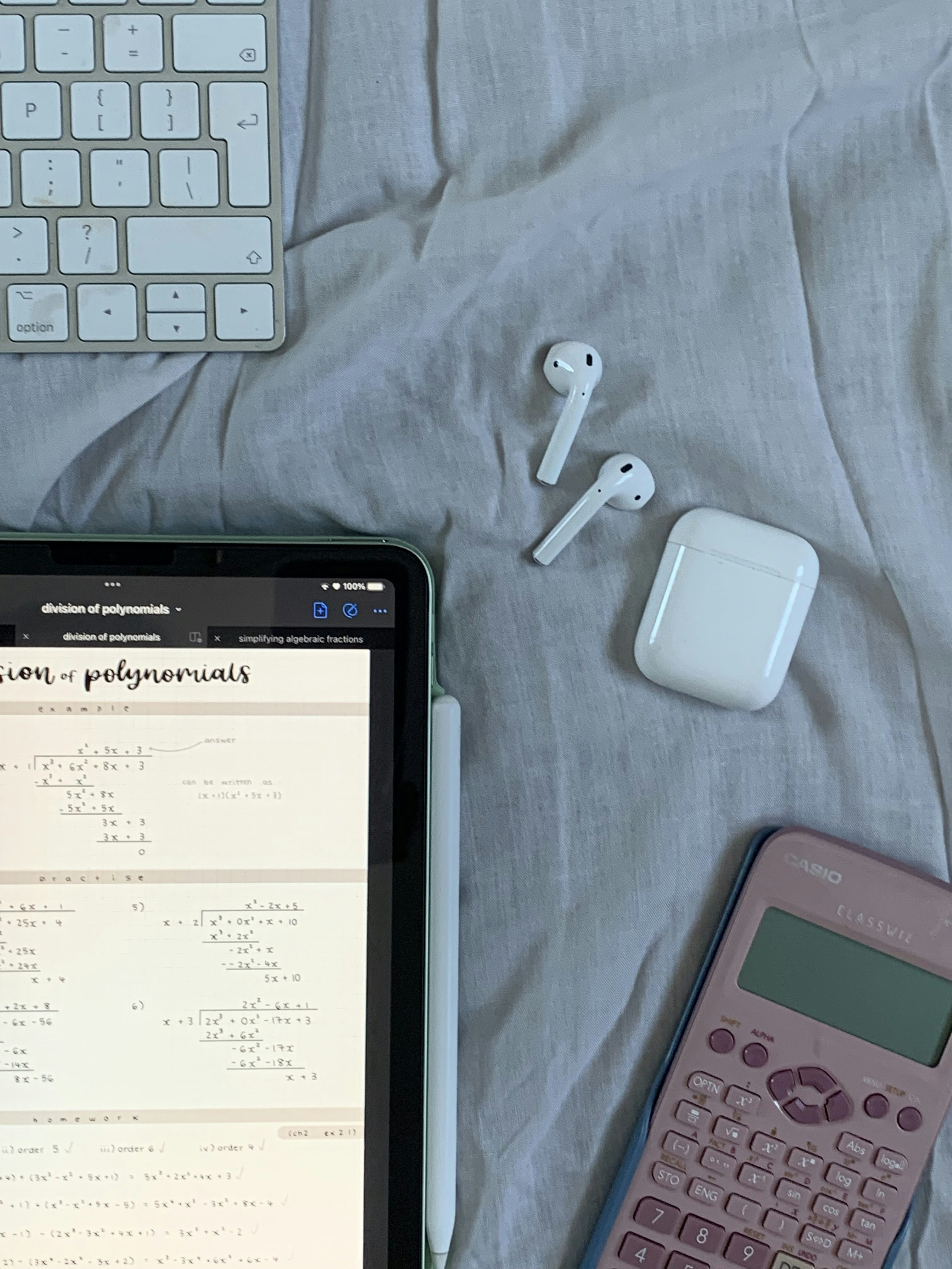 A tablet displaying mathematical problems is placed on a cloth surface. Nearby are a white stylus, a wireless keyboard, a pair of white wireless earbuds with their case, and a pink calculator.