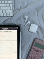 A tablet displaying mathematical problems is placed on a cloth surface. Nearby are a white stylus, a wireless keyboard, a pair of white wireless earbuds with their case, and a pink calculator.