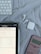 A tablet displaying mathematical problems is placed on a cloth surface. Nearby are a white stylus, a wireless keyboard, a pair of white wireless earbuds with their case, and a pink calculator.