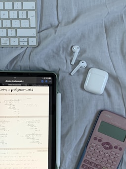 A tablet displaying mathematical problems is placed on a cloth surface. Nearby are a white stylus, a wireless keyboard, a pair of white wireless earbuds with their case, and a pink calculator.