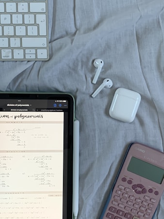A tablet displaying mathematical problems is placed on a cloth surface. Nearby are a white stylus, a wireless keyboard, a pair of white wireless earbuds with their case, and a pink calculator.