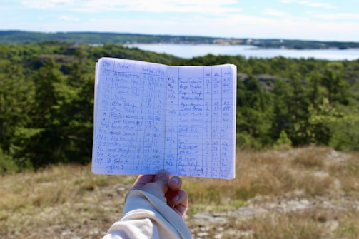 A person holds an open notebook filled with handwritten notes and numbers. The background features a scenic view of a landscape with dense green trees, a body of water, and a partly cloudy sky.