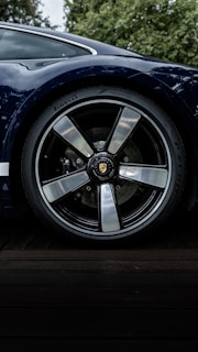 A close-up shot of a car wheel featuring a sleek, polished rim and a high-performance tire branded with Pirelli. The car's bodywork, in a glossy dark blue, is partially visible, reflecting the surrounding greenery.