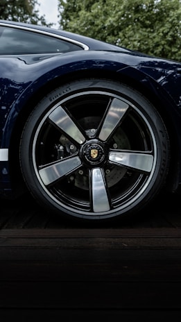 A close-up shot of a car wheel featuring a sleek, polished rim and a high-performance tire branded with Pirelli. The car's bodywork, in a glossy dark blue, is partially visible, reflecting the surrounding greenery.