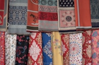 A collection of colorful fabrics is displayed in layers. The fabrics feature intricate patterns, including floral designs, geometric shapes, and traditional motifs. The textures vary, adding depth and richness to the display.