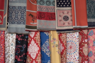 A collection of colorful fabrics is displayed in layers. The fabrics feature intricate patterns, including floral designs, geometric shapes, and traditional motifs. The textures vary, adding depth and richness to the display.