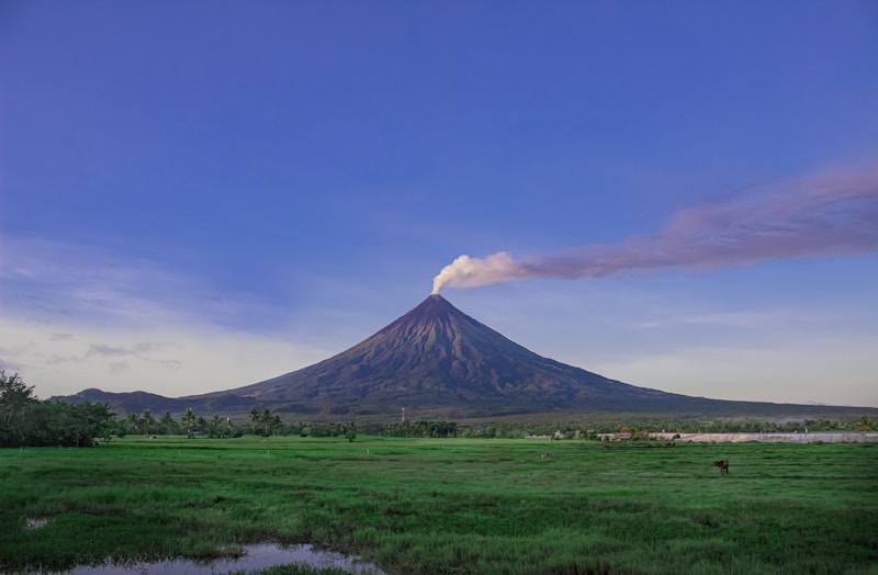 A majestic volcano rises from a lush green landscape, with a wisp of smoke emanating from the crater under a clear blue sky. The vibrant greenery contrasts with the dark grey slopes of the volcano, providing a serene and picturesque scene.