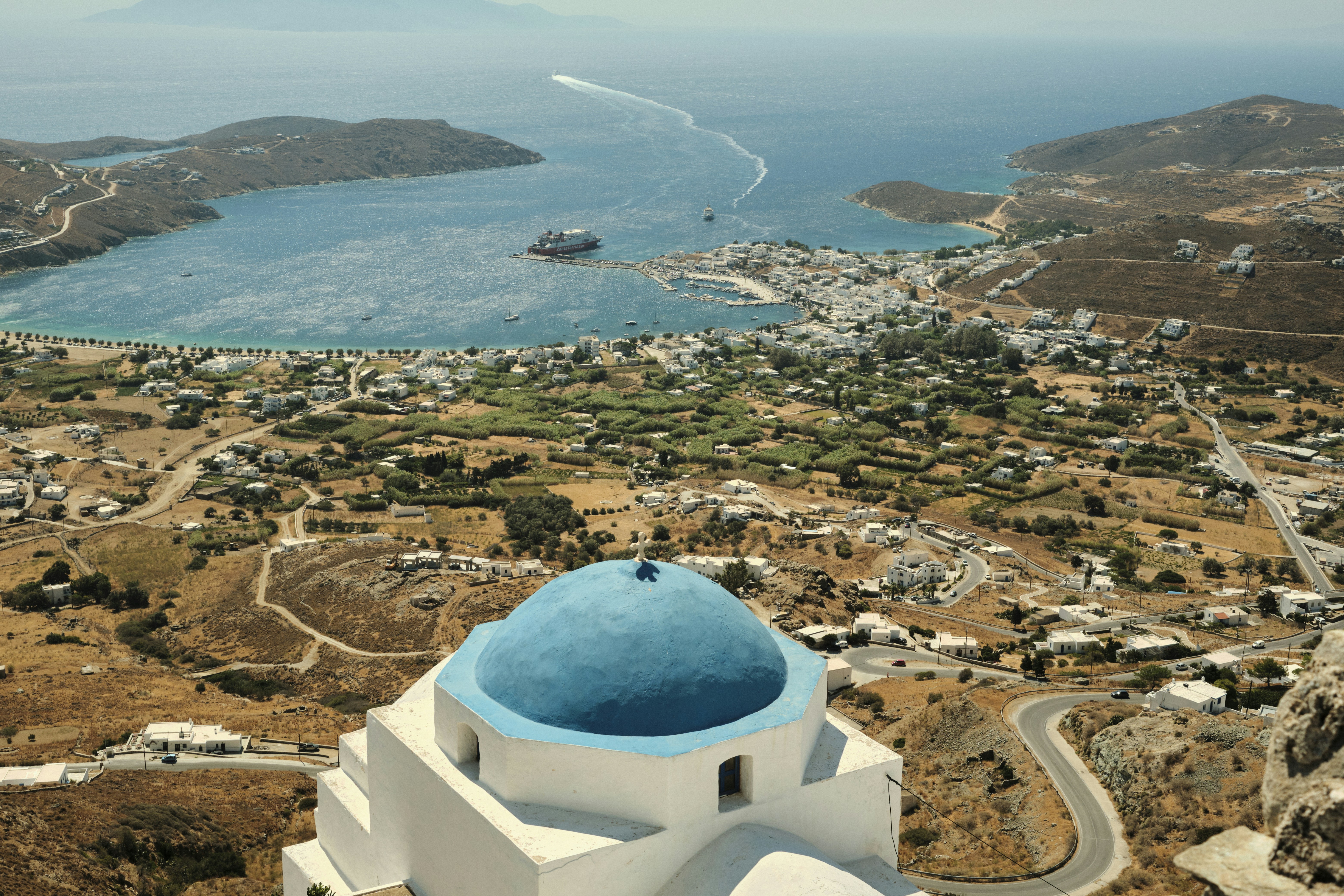 a white building with a blue dome and a blue dome on a hill overlooking a city and water, View of the port of Serifos and its surroundings from the Chora
