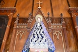 A religious statue of a figure draped in an ornate blue robe with intricate white decorations, standing in front of an elaborate wooden altar backdrop. The altar features Gothic-style architectural elements and a prominent wooden cross at the top.