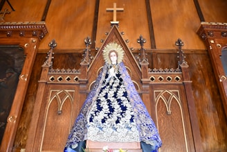 A religious statue of a figure draped in an ornate blue robe with intricate white decorations, standing in front of an elaborate wooden altar backdrop. The altar features Gothic-style architectural elements and a prominent wooden cross at the top.