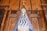 A religious statue of a figure draped in an ornate blue robe with intricate white decorations, standing in front of an elaborate wooden altar backdrop. The altar features Gothic-style architectural elements and a prominent wooden cross at the top.