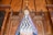 A religious statue of a figure draped in an ornate blue robe with intricate white decorations, standing in front of an elaborate wooden altar backdrop. The altar features Gothic-style architectural elements and a prominent wooden cross at the top.