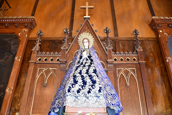 A religious statue of a figure draped in an ornate blue robe with intricate white decorations, standing in front of an elaborate wooden altar backdrop. The altar features Gothic-style architectural elements and a prominent wooden cross at the top.