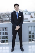 A young man dressed in a formal suit, complete with a white shirt and blue tie, stands on a rooftop with a cityscape in the background. He is facing forward with his hands clasped in front of him, displaying a name tag on his jacket.