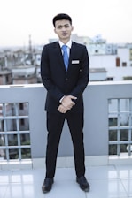 A young man dressed in a formal suit, complete with a white shirt and blue tie, stands on a rooftop with a cityscape in the background. He is facing forward with his hands clasped in front of him, displaying a name tag on his jacket.