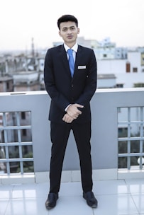 A young man dressed in a formal suit, complete with a white shirt and blue tie, stands on a rooftop with a cityscape in the background. He is facing forward with his hands clasped in front of him, displaying a name tag on his jacket.