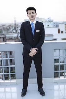 A young man dressed in a formal suit, complete with a white shirt and blue tie, stands on a rooftop with a cityscape in the background. He is facing forward with his hands clasped in front of him, displaying a name tag on his jacket.