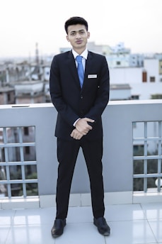 A young man dressed in a formal suit, complete with a white shirt and blue tie, stands on a rooftop with a cityscape in the background. He is facing forward with his hands clasped in front of him, displaying a name tag on his jacket.