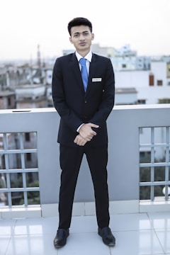 A young man dressed in a formal suit, complete with a white shirt and blue tie, stands on a rooftop with a cityscape in the background. He is facing forward with his hands clasped in front of him, displaying a name tag on his jacket.