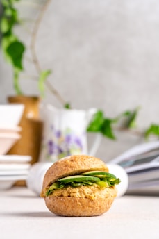 A single bread roll sandwich is placed in a light, airy setting. The sandwich is filled with a mixture that appears to be a spread or filling with green leaves, possibly lettuce or spinach. In the background, there is a soft-focused arrangement of stacked dishes, a decorative jug with floral patterns, and an out-of-focus green plant.