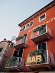 A red building with multiple balconies adorned with green plants. Signs for 'HOTEL' and 'BAR' are prominently displayed on the facade. The building features dark green shutters and architectural details reminiscent of European style.